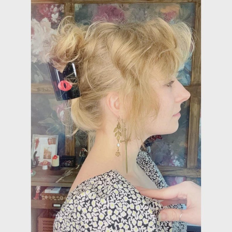 May include: A black hair clip with a red eye design is placed in blonde hair. The person is wearing a floral print top and gold-colored leaf earrings. The background includes a window and a shelf with various items.