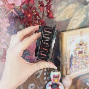May include: A black rectangular hair claw clip with a wavy design. The clip is held in a hand, with a blurred background of flowers and artwork.