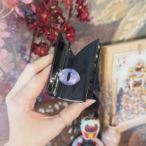 May include: Black claw hair clip with a cat eye design. The clip is black with a purple and white eye-shaped embellishment. The clip is held in a hand, with a silver ring on the finger.
