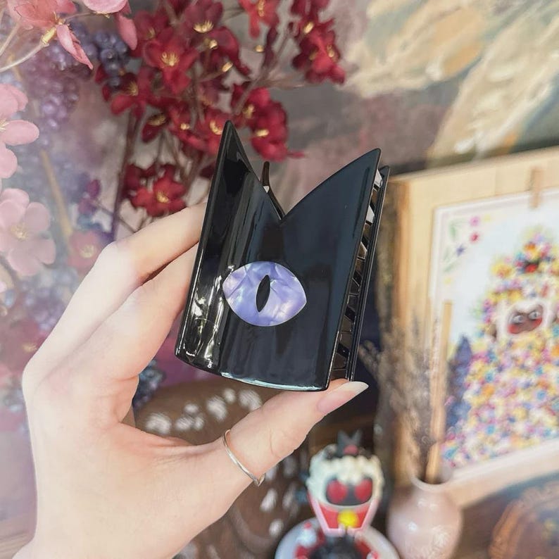 May include: Black claw hair clip with a cat eye design. The clip is black with a purple and white eye-shaped embellishment. The clip is held in a hand, with a silver ring on the finger.