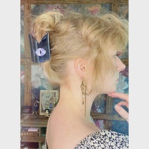 May include: A black hair clip with a purple eye design is placed in blonde hair. The clip has a geometric shape and a glossy finish. Gold-colored earrings with leaf and pearl accents are also visible.