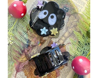 May include: A black hair claw clip featuring two large oval eyes with light blue irises and black pupils, accented with small purple, light blue, and yellow star-shaped embellishments on the surface and a black plastic spring-loaded mechanism.