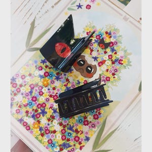 May include: Two black hair claw clips with a glossy finish. One clip features a red eye design, and the other is a classic rectangular shape. The clips are displayed on a floral patterned background.