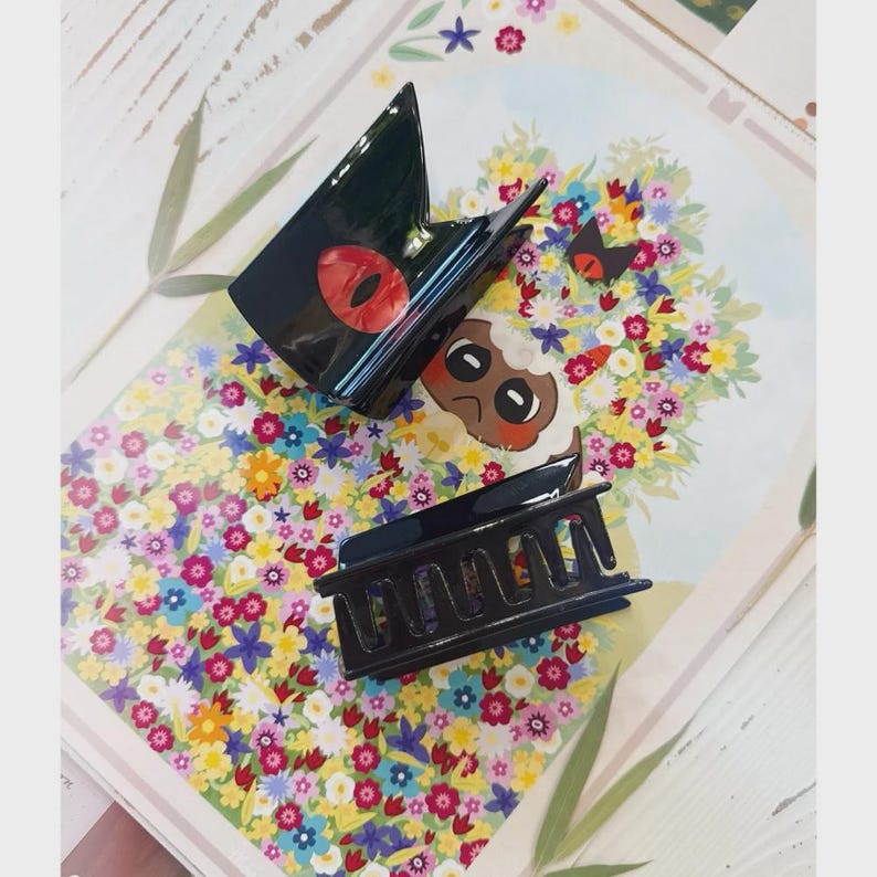 May include: Two black hair claw clips with a glossy finish. One clip features a red eye design, and the other is a classic rectangular shape. The clips are displayed on a floral patterned background.