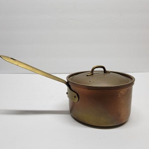 May include: A copper saucepan with a brass handle and lid. The pot has a rounded body and a long, flat handle extending from the side. The lid has a small loop handle. The copper has a warm, reddish-brown hue, with brass accents.