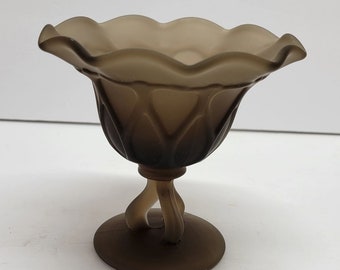 May include: A frosted brown glass pedestal bowl with a scalloped rim. The bowl has a tulip-shaped design with a twisted stem and a circular base. The glass has a matte finish.