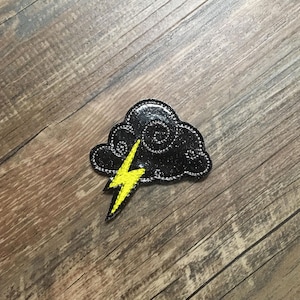 May include: A black and yellow embroidered patch of a cloud with a lightning bolt. The patch is on a woodgrain background.