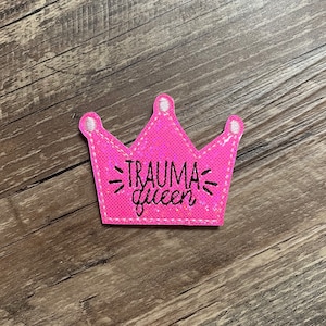 May include: Pink felt crown with the text "TRAUMA queen" in black lettering.