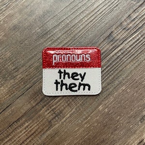 以下が含まれることがあります： 白と赤のグリッターパッチに、黒い刺繍で「pronouns they them」と書いてあります。