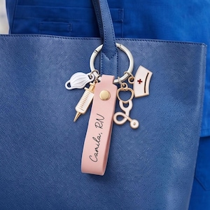May include: A navy blue tote bag with a pink leather keychain. The keychain features medical-themed charms, such as a syringe, stethoscope, and face mask. The keychain also has the text "Camila, RN".