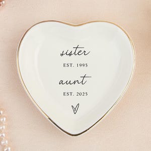 以下が含まれることがあります： 金色の縁取りが付いた、ハート型の白いセラミック製の小物入れ。 黒い筆記体で「sister」と「aunt」の文字が書かれており、それぞれの文字の下に「EST. 1995」と「EST. 2025」の年号が書かれています。 下部には小さなハートのシンボルがあります。