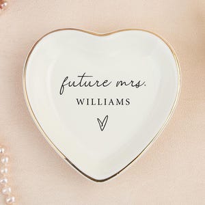 Peut inclure: Un vide-poches en céramique blanc en forme de cœur, avec un bord doré. Le plat porte l'inscription "future mrs. WILLIAMS" en écriture noire, avec un petit symbole de cœur en dessous. Un cadeau de fiançailles ou de mariage.