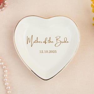May include: A heart-shaped, off-white trinket dish with a gold rim. The dish has the words "Mother of the Bride" and the date 12.10.2025 in gold script. The background is a light pink surface with floral and pearl accents.