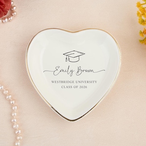 May include: Heart-shaped, off-white ceramic trinket dish with a gold rim. The dish features a graduation cap graphic and the text "Emily Brown, Westbridge University, Class of 2026". A decorative item for a graduate.