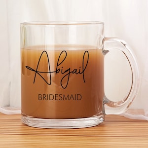 May include: A clear glass mug filled with a light brown liquid, featuring the name "Abigail" in a stylish script and the word "BRIDESMAID" below. The mug has a curved handle and is set against a soft, blurred background.