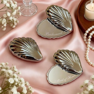 Op de afbeelding: Zilveren schelpvormige compacte spiegels met de woorden "Bridesmaid" en "Maid of Honor" gegraveerd. De spiegels liggen op een roze satijnen oppervlak, met witte bloemen, een kaars en een parelketting.