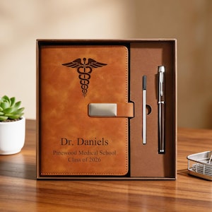 Può includere: Un set regalo con diario e penna in similpelle marrone. Il diario presenta un simbolo medico del caduceo e il nome "Dr. Daniels" con "Pinewood Medical School Class of 2026". Include una penna argentata e una penna nera.