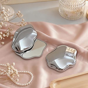 May include: Two silver-colored, organically shaped compact mirrors are displayed on a pink satin surface. One mirror is open, revealing a reflective surface. The other is closed. A pearl necklace and decorative flowers are also present.