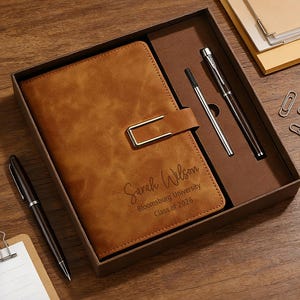 May include: A brown leather journal with a metal clasp, personalised with the name "Sarah Wilson" and "Bloomsburg University Class of 2026." The set includes two pens, paperclips, and a notepad, all presented in a gift box.