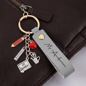 Könnte beinhalten: Ein grauer Leder-Schlüsselanhänger mit dem Namen "Mr. Anderson" und goldfarbenen Elementen. Der Schlüsselanhänger hat Anhänger von einem roten Apfel, einem Bleistift, einem Lineal und einem Schild mit der Aufschrift "#1 Teacher".