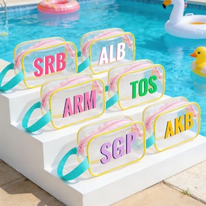 Puede incluir: Bolsas de cosméticos rectangulares transparentes con iniciales coloridas. Cada bolsa tiene una cremallera rosa, ribete amarillo y asa turquesa. Las bolsas están dispuestas sobre escalones blancos junto a una piscina.