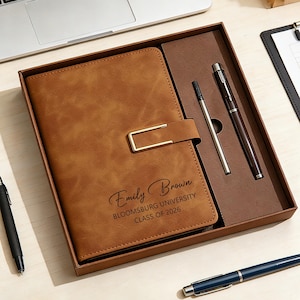 Op de afbeelding: Een bruin leren notitieboek met een gouden sluiting, gepersonaliseerd met de naam "Emily Brown" en "Bloomsburg University Class of 2026." Het notitieboek zit in een geschenkdoos met twee pennen. Een zwarte pen en een blauwe pen zijn ook zichtbaar.