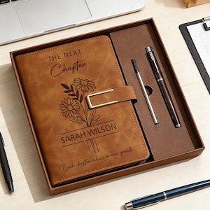 Puede incluir: Un diario de cuero marrón con las palabras "The Next Chapter" y "Sarah Wilson" grabadas en la portada, junto con un diseño floral. El diario está en una caja de regalo con dos bolígrafos. También se ve un bolígrafo negro y un bolígrafo azul.