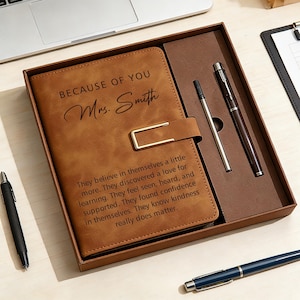May include: A brown leather journal set in a gift box. The journal has the text "BECAUSE OF YOU Mrs. Smith" and an inspirational quote. The set includes two pens and a pen holder. A blue pen is also visible.