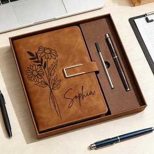 May include: A brown leather journal with a floral design and the name "Sophia" engraved on the cover. The journal is in a gift box with two pens. One pen is black, and the other is brown.
