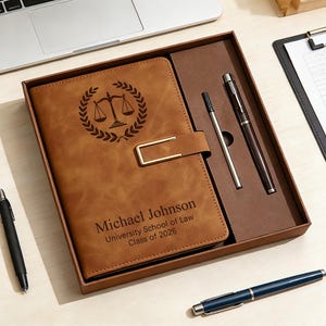 Può includere: Un diario in pelle marrone con chiusura dorata, un'emblema della bilancia della giustizia e testo personalizzato. Il diario è in una confezione regalo con due penne. Sono visibili anche una penna nera e una blu.