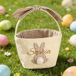 以下が含まれることがあります： 内側とハンドルのリボンに茶色のギンガムチェック模様が入った、クリーム色のイースターバスケット。バスケットには刺繍されたウサギのデザインと「Sophia」という名前が施されています。バスケットは、カラフルなイースターエッグが置かれた緑の草の上にあります。