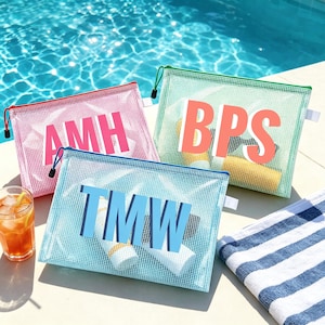 May include: Three mesh zipper pouches in pink, green, and blue, each with a different set of initials in a bold font. The pink pouch has red zipper, the green pouch has a green zipper, and the blue pouch has a blue zipper. The pouches are near a pool.