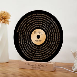 May include: A black vinyl record with gold lettering, featuring a song lyric about love and a place for someone in your life. The record is on a wooden stand with the text "Bailey & David 10.02.2025".