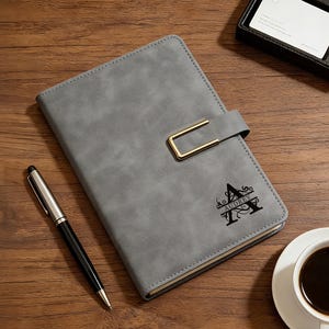 Op de afbeelding: Grijze kunstleren journal met een goudkleurige sluiting en de naam "Audrey" en de letter "A" op de kaft gedrukt. Een zilveren en zwarte pen en een kop koffie liggen naast het journal op een houten oppervlak.