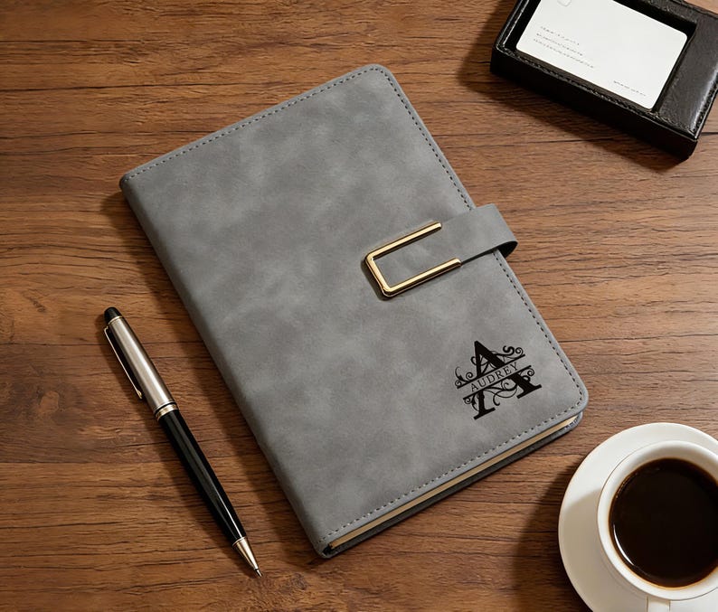 Op de afbeelding: Grijze kunstleren journal met een goudkleurige sluiting en de naam "Audrey" en de letter "A" op de kaft gedrukt. Een zilveren en zwarte pen en een kop koffie liggen naast het journal op een houten oppervlak.