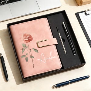 Op de afbeelding: Een roze notitieboek met een roosillustratie en de naam Amelia in wit handschrift, afgesloten met een goudkleurige metalen sluiting. Het notitieboek wordt gepresenteerd in een zwarte geschenkdoos naast een zwarte pen en een zilverkleurige vulling.