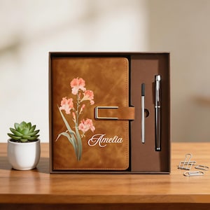 May include: A brown leather journal with a floral design and the name "Amelia" is displayed in a gift box with a pen and a pen refill. A small succulent in a white pot is on the left.