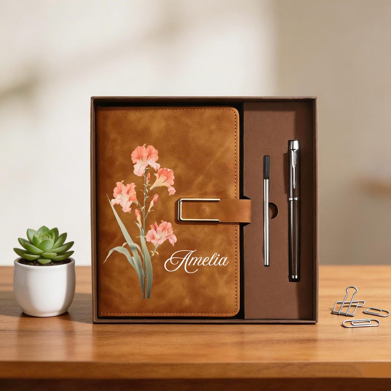 May include: A brown leather journal with a floral design and the name "Amelia" is displayed in a gift box with a pen and a pen refill. A small succulent in a white pot is on the left.