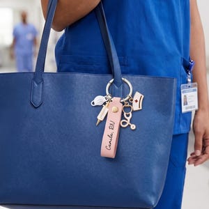 Pu&ograve; includere: Una borsa tote blu navy con un portachiavi in pelle rosa. Il portachiavi presenta ciondoli a tema medico, tra cui una siringa, uno stetoscopio e una mascherina. Il portachiavi riporta anche la scritta "Camila, RN".