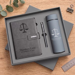 Op de afbeelding: Een grijze geschenkset in een doos met een notitieboek met een weegschaal van justitie en de tekst Theodore Lane Attorneys at Law, een bijpassende metalen pen en een grijze cilindrische isolatiefles met hetzelfde icoon en tekst erop.