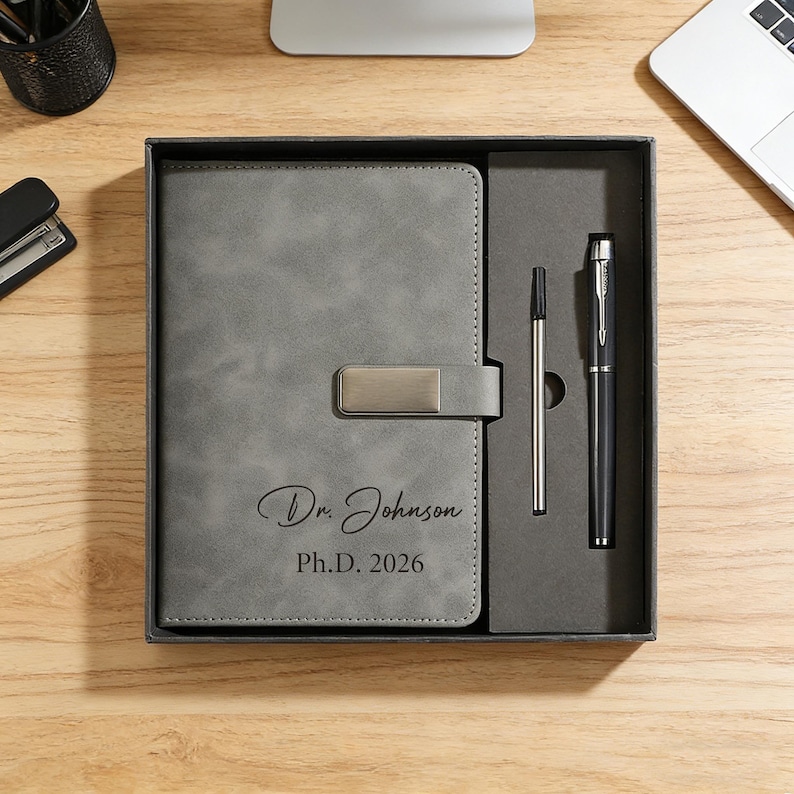 May include: A gray journal and pen set in a gift box. The journal has a metal clasp and the inscription "Dr. Johnson, Ph.D. 2026". The set includes a silver pen and a black pen.