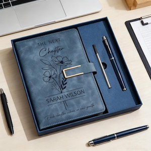 K&ouml;nnte beinhalten: Ein blaues Notizbuch-Set aus Kunstleder mit passendem Stift und einem silbernen Stift. Das Notizbuchcover tr&auml;gt die Aufschrift "THE NEXT Chapter" und "SARAH WILSON" mit einem floralen Design. Das Notizbuch befindet sich in einer blauen Geschenkbox.
