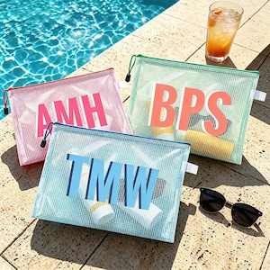 May include: Three mesh zipper pouches in pink, green, and blue, each with a different set of initials in bold letters. The pink pouch has "AMH", the green pouch has "BPS", and the blue pouch has "TMW". Sunglasses and a drink are next to the pouches.