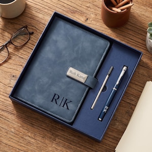 May include: A blue notebook with a magnetic closure featuring a silver plate engraved with the name Rob Kane and the initials R|K embossed on the cover. The set includes a matching blue pen with the initials R|K and a silver ink refill cartridge.