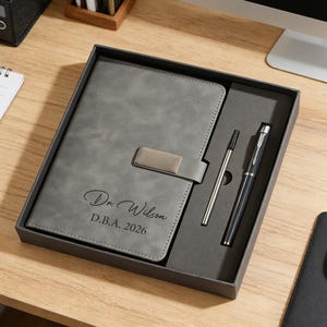 May include: A gray notebook and pen set in a gift box. The notebook has a silver clasp and the name "Dr. Wilson" and "D.B.A. 2026" printed on the cover. The set includes a silver pen and a black pen.
