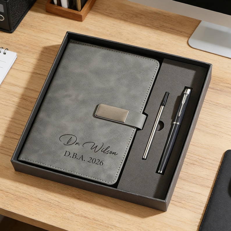 May include: A gray notebook and pen set in a gift box. The notebook has a silver clasp and the name "Dr. Wilson" and "D.B.A. 2026" printed on the cover. The set includes a silver pen and a black pen.