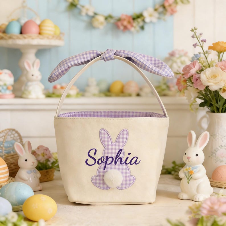 Pu&ograve; includere: Cestino di Pasqua color crema con un'applicazione a coniglietto a quadretti viola e il nome "Sophia" ricamato in viola. Il cestino ha un manico e un interno a quadretti abbinati. &Egrave; attaccata una soffice coda a pompon bianca. Uova di Pasqua e coniglietti in ceramica circondano il cestino.