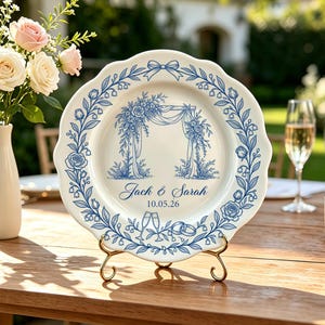 Pode incluir: Um prato de cerâmica branca com borda recortada, apresentando uma borda floral azul e uma ilustração central de um arco de casamento. O texto "Jack & Sarah" e "10.05.26" está impresso abaixo do arco, com duas taças e alianças.