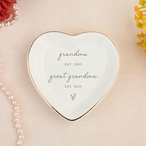 May include: A white heart-shaped ceramic trinket dish with a gold-colored rim. The center features black text reading grandma EST. 1995, great grandma EST. 2025, and a small hand-drawn heart symbol at the bottom of the dish.