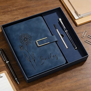 Puede incluir: Un juego de diario de cuero azul con un dise&ntilde;o floral y el nombre "Bailey" grabado en la portada. El juego incluye dos bol&iacute;grafos, un portal&aacute;pices y clips, todo presentado en una caja azul oscuro.